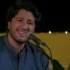 A smiling male singer performing live at night with a microphone, dressed in traditional attire, with soft lights glowing in the background