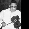 A black-and-white photograph of an Indian classical violinist seated and playing the violin, wearing traditional attire, with a focused expression against a patterned backdrop.