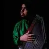 A man in a green kurta holding a string instrument, looking sideways against a dark background.
