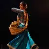 A Kathak dancer in a blue costume twirling gracefully on stage under warm lighting.
