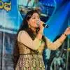 A female vocalist performing on stage, singing into a handheld microphone with expressive hand gestures, dressed in a beige and black embroidered outfit with gold accessories, standing beside a music stand against a lit stage backdrop.