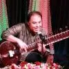 An Indian classical musician seated on stage is playing a sitar while smiling and speaking into a microphone, wearing a patterned kurta, with decorative lights and floral arrangements in the background.