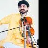 A violinist seated and performing, holding a violin and bow, wearing a yellow patterned kurta and glasses, positioned in front of a microphone against a plain light background.
