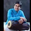A mridangam artist seated with the instrument placed horizontally across his lap, wearing a blue kurta and white attire, looking at the camera against a dark studio background.