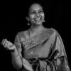 A black-and-white portrait of a smiling woman wearing a traditional silk saree and jewelry, gesturing with one hand as if explaining or performing, set against a plain dark background.