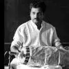 A male mridangam artist dressed in traditional attire sits on stage holding a mridangam, poised to perform during an Indian classical music concert.
