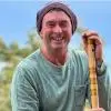 Man holding a bansuri (bamboo flute) outdoors, smiling at the camera, wearing a headscarf and casual clothing with a blurred natural background.