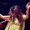 Color photograph of a female performer on stage, captured mid-performance with both arms raised in an expressive gesture. She is wearing a sleeveless, patterned outfit with bangles on her wrists. A microphone is positioned above her, and a guitarist is visible in the background. The stage lighting is warm and focused, emphasizing movement and musical energy.