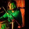 A tabla player smiling during a live performance, seated behind the tabla with microphones around the instrument under colorful stage lighting.