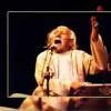 Color photograph of a male singer performing on stage, captured in a powerful, emotional moment with his mouth open mid-song and one hand raised expressively. He is seated behind a harmonium, singing into a microphone. The lighting is warm and focused, and the image has a framed or bordered presentation.