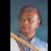 Portrait of bansuri maestro Pt. Venkatesh Godkhindi holding a bamboo flute, wearing a blue kurta, against a plain background.