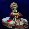 Kathakali performer Ramyani Roy in elaborate traditional costume and green facial makeup performing on stage during a classical Kathakali dance presentation.