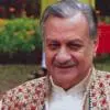 Color portrait of an elderly man with neatly combed grey hair and a moustache, smiling gently. He is wearing a traditional outfit with a floral-patterned vest. The background features a garden setting with greenery and flowers, softly out of focus.