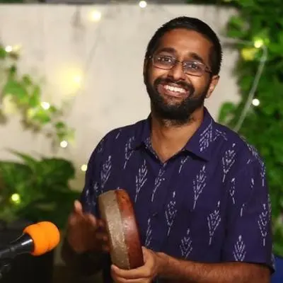 Percussion artist holding a frame drum during a live performance with greenery backdrop.