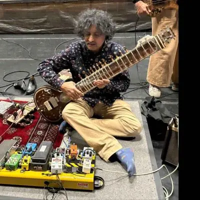 An Indian classical musician with curly hair is seated cross-legged on stage, playing a sitar, surrounded by multiple electronic effects pedals and cables, with other instruments and equipment visible nearby.