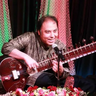 An Indian classical musician seated on stage is playing a sitar while smiling and speaking into a microphone, wearing a patterned kurta, with decorative lights and floral arrangements in the background.