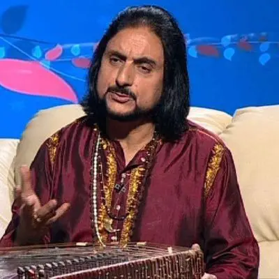 An Indian classical musician wearing a maroon kurta is seated on a sofa, holding a santoor and gesturing while speaking during a studio interview, with a blue decorative backdrop behind him.