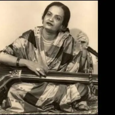 A black-and-white photograph of a female Indian classical musician seated cross-legged, playing a veena, wearing a traditional saree and jewelry, with a thoughtful expression against a plain background.