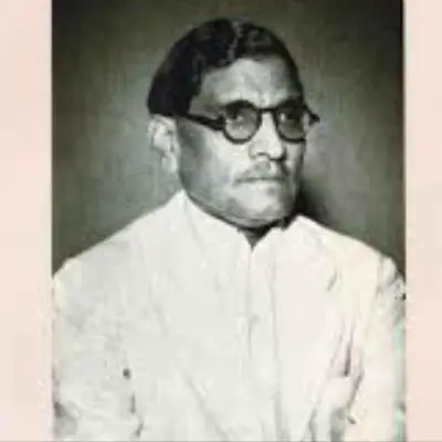 A black-and-white portrait of an Indian classical musician wearing glasses and traditional attire, looking to the side with a composed expression against a plain background.