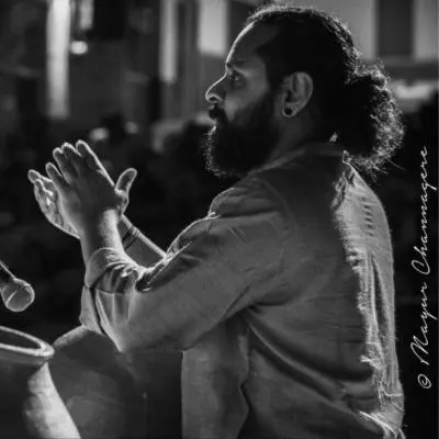 A male ghatam artist with tied-back hair and beard performs on stage, clapping rhythmically beside a clay pot (ghatam) with a microphone placed nearby during a live Indian classical concert.