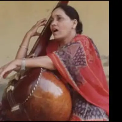 Color photograph of a female classical musician seated with a tanpura, holding the instrument upright while singing. She is wearing a traditional saree with a red shawl draped over her shoulder. Her expression is engaged and expressive, suggesting a live or practice performance setting.