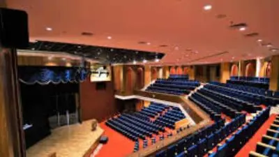 Interior view of the main auditorium at Rajasthan International Centre, featuring tiered blue seating, a stage with curtains, and warm lighting.