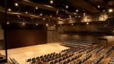 An interior view of the Experimental Theatre at NCPA, Mumbai. It shows a versatile, black-box style performance space with a polished light-wood stage area. The seating is arranged in a tiered, semi-circular fashion with dark chairs on wooden platforms. The ceiling features exposed industrial pipes and professional stage lighting fixtures distributed across the hall.