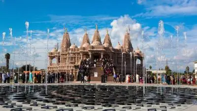 Exterior view of BAPS Swaminarayan Temple featuring a large intricately carved stone structure with multiple domes and spires, a central staircase with visitors gathered, and a foreground plaza with decorative water fountains under a clear blue sky.