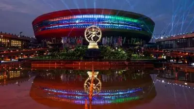 Night view of Bharat Mandapam in New Delhi featuring colorful architectural lighting, the iconic Nataraja statue, and reflections in the water foreground.