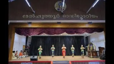 Classical dance performance at Melpathur Auditorium featuring Bharatanatyam dancers on stage with live musicians seated to the side beneath the auditorium signage.