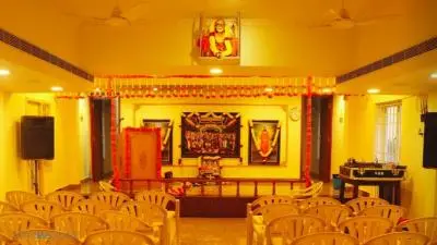Interior view of Sri Krishna Saras Hall featuring a small decorated stage, devotional portraits, and rows of seating arranged for a cultural or spiritual gathering.
