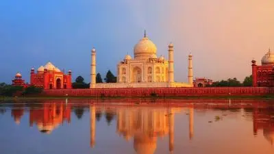 Scenic view of the Taj Mahal at sunrise, with its white marble dome and minarets reflected in calm water, surrounded by red sandstone buildings under a colorful sky.