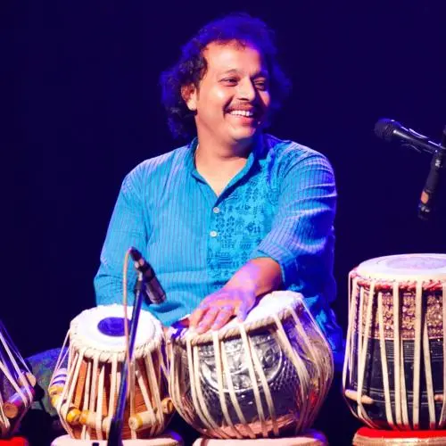 Tabla musician seated on stage surrounded by Indian percussion drums and microphones.