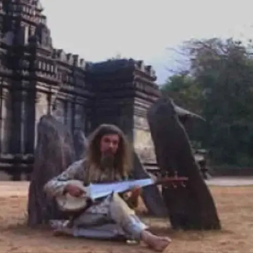 A long-haired musician seated cross-legged outdoors, playing a string instrument in front of ancient stone temple structures, surrounded by large upright stones in a rustic setting.