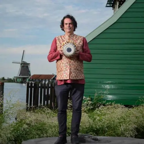 A man standing outdoors near a waterfront, holding a tabla drum, with a green wooden house and a windmill visible in the background.