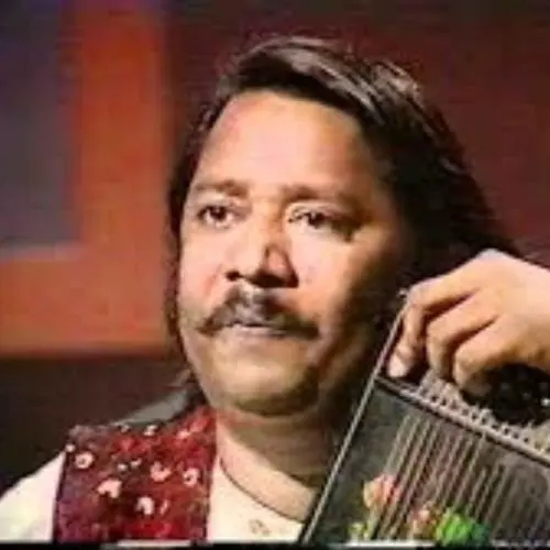 A middle-aged Indian classical vocalist with long hair and a moustache is seated, holding a swarmandal, wearing a patterned shawl, and looking attentively during a performance or interview setting.