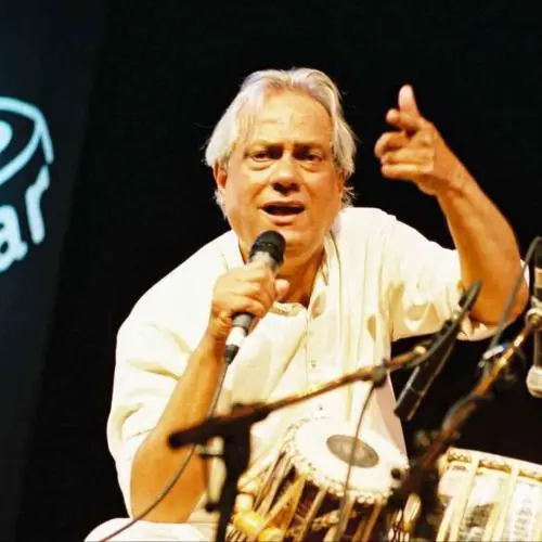 An Indian classical tabla player performing on stage, holding a microphone and gesturing with one hand while seated behind the tabla, under warm stage lighting against a dark background.