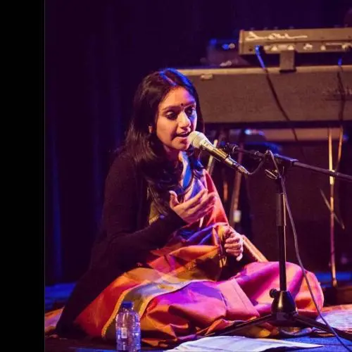 A female vocalist seated cross-legged on stage, singing into a microphone, wearing a colorful saree, with a keyboard setup visible in the background under warm stage lighting.