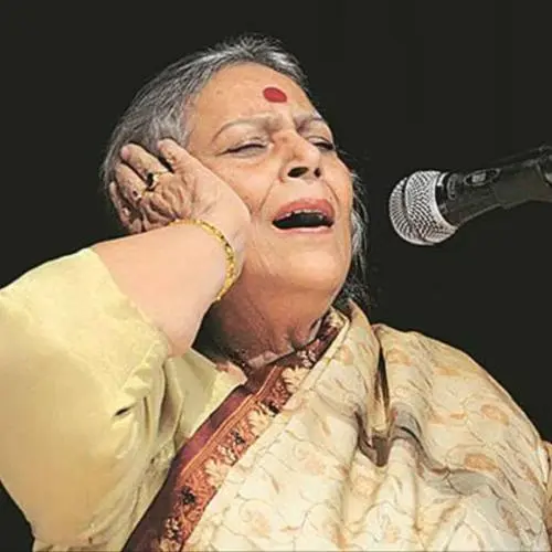 An elderly Indian classical vocalist wearing a saree and a large red bindi, singing into a microphone with eyes closed and hand placed on her ear, conveying deep emotion during a live performance.