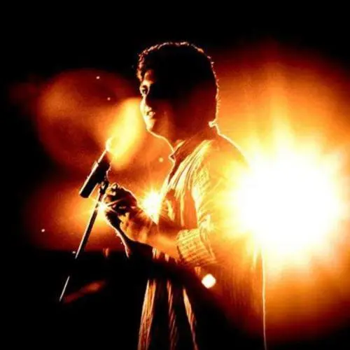 A vocalist singing into a microphone on stage, illuminated by bright backlighting during a live performance.