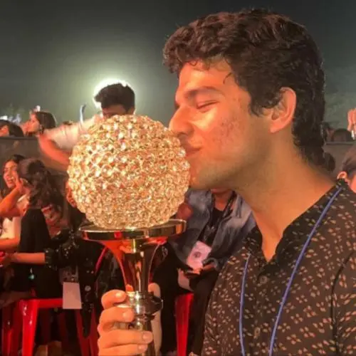 A young man in a patterned black kurta holds and kisses a large decorative trophy with a crystal-like globe top, standing in a crowded outdoor event at night with bright stage lights and seated audience members visible in the background.