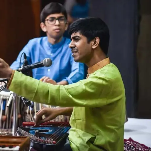 An Indian classical vocalist in a green kurta sings into a microphone while playing a swarmandal, gesturing expressively with one hand; a tabla player in a blue kurta accompanies in the background.
