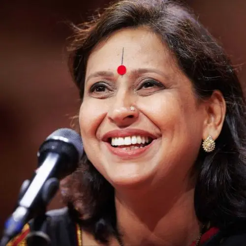 Indian classical vocalist performing on stage, smiling while singing into a microphone, wearing a red bindi and traditional earrings against a warm concert hall background.