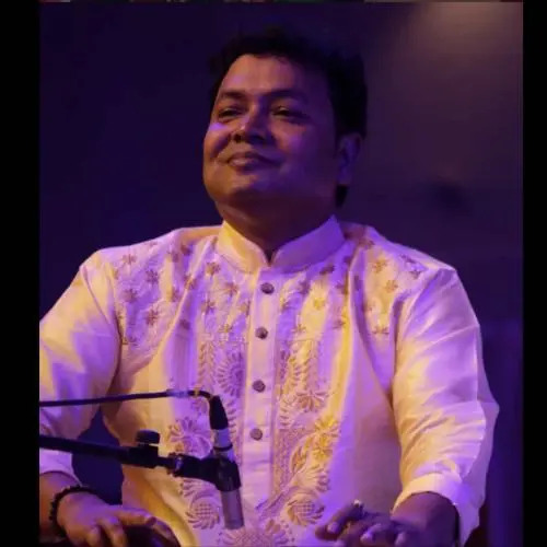 An Indian classical musician in a cream embroidered kurta sits on stage under purple lighting, smiling gently while performing, with a microphone positioned in front of him.