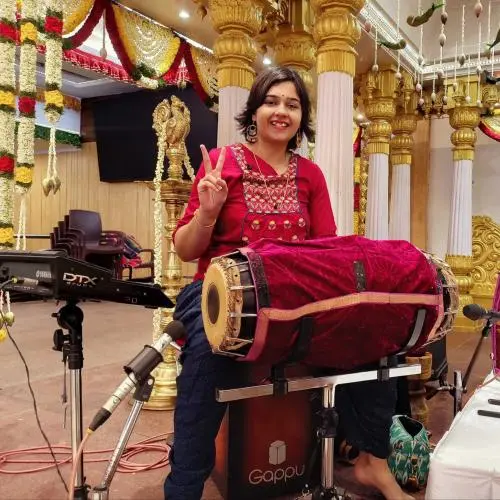 Aswini Srinivasan seated on stage with a mridangam during a Carnatic music performance in a decorated temple hall.