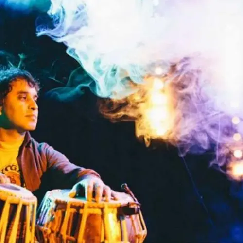 Bobby Singh performing tabla on stage, seated with hands on the drums, illuminated by colorful stage lights and surrounded by atmospheric smoke effects.