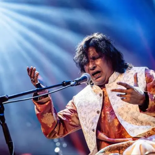 Color photograph of a male singer performing on stage, captured mid-expression with eyes closed and hands raised in an emotive gesture. He is seated and singing into a microphone, wearing a richly embroidered traditional outfit. Dramatic stage lighting with beams of light from above enhances the intensity of the performance.