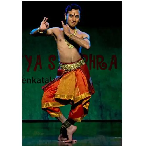 Male Bharatanatyam dancer performing on stage, wearing a traditional orange and gold costume with temple jewellery, holding a balanced pose on one leg with expressive hand mudras against a dark green backdrop.