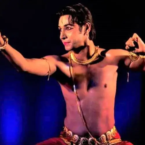 Male Indian classical dancer performing on stage, wearing traditional jewelry and a red costume, striking a dramatic dance pose with extended arms under blue stage lighting.