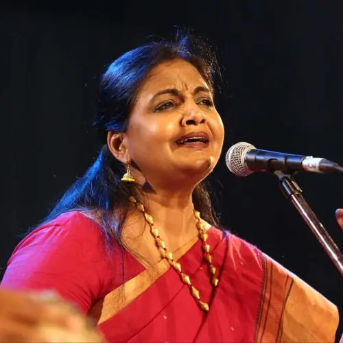 A female Carnatic vocalist in a red saree sings expressively into a microphone on stage, with a raised hand gesture during a live classical music performance.